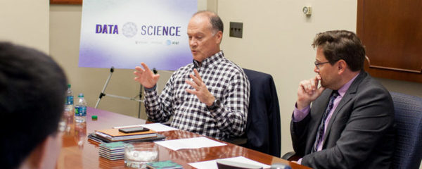 John Donovan and Elliot Visconsi sit at a meeting table having a discussion with a Data Science placard in the background.