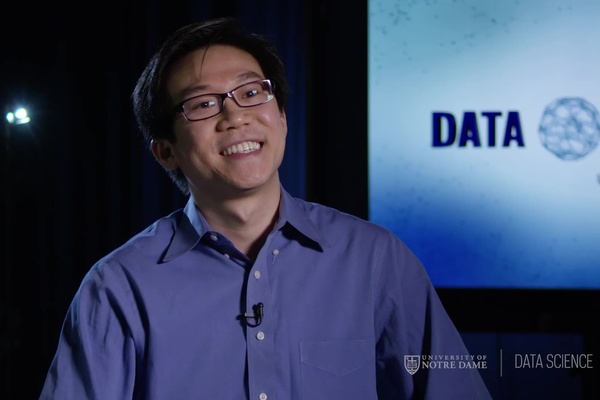 Huy Huynh in a dark blue button down shirt smiles in front of a digital screen during production of an instructional video.