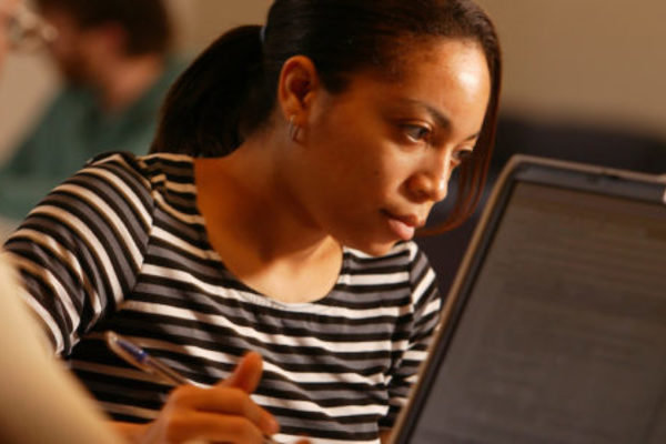 A woman looks at a document while another person looks at a laptop screen in the foreground.