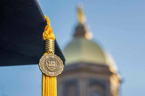 Graduation cap with gold tassel and Notre Dame emblem with a blurred golden dome in the background.