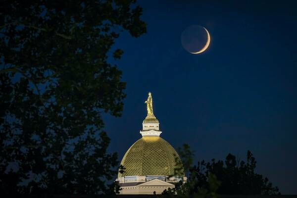 Night time picture of Notre Dame's golden dome centered with foliage on the left and a crescent moon to the top right.