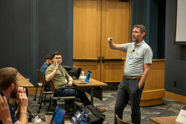 A professor standing at the front of a room pointing to the audience of students sitting at desks.